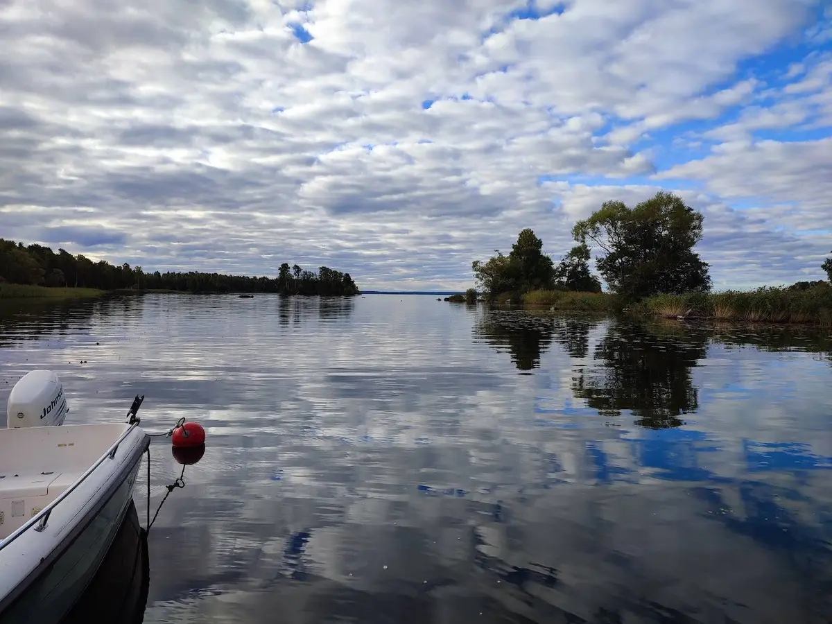 En solig sommardag vid Herrfallet AB med blå himmel och moln som speglas i det lugna vattnet.