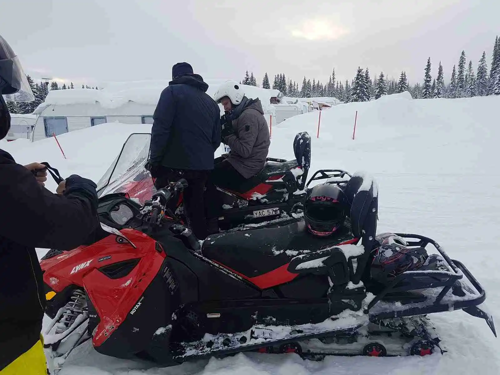 En vinterlandskap med snöskoteråkare i Åkersjöns sportcamping. Snön täcker marken och omgivningen, medan personer utrustade med skyddsutrustning kör snöskotrar. En isig och kylig atmosfär omger platsen, perfekt för vintersporter.