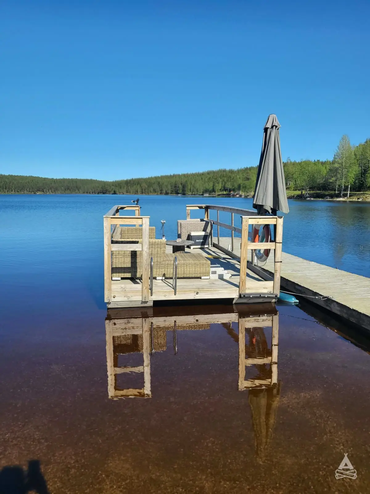 En bild av en brygga som sträcker sig ut över en fridfull sjö vid Rödvattnet camping. Omkring bryggan breder det stilla vattnet ut sig, omgiven av gröna träd och skog.