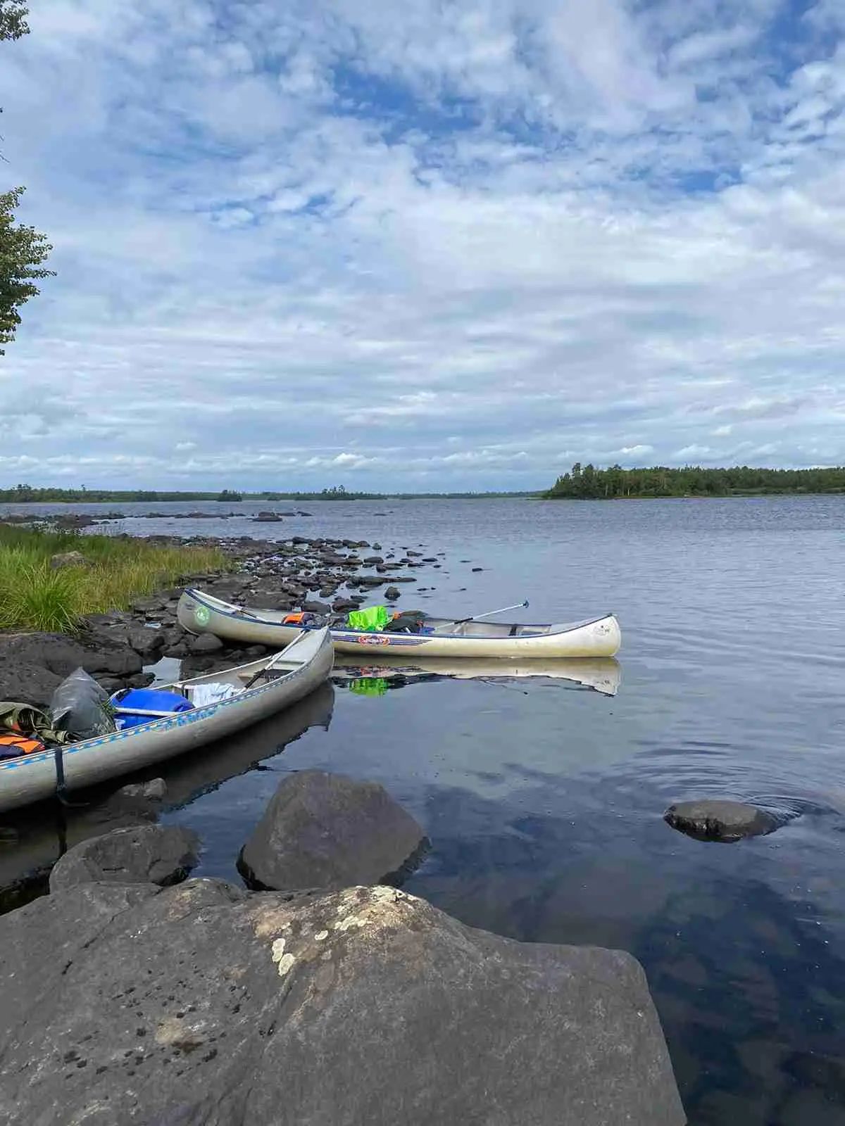 En solig dag vid Agundaborg Camp & Paddle, där flera kajaker ligger förtöjda vid en sjö omgiven av grönskande landskap och en blå himmel med några moln.