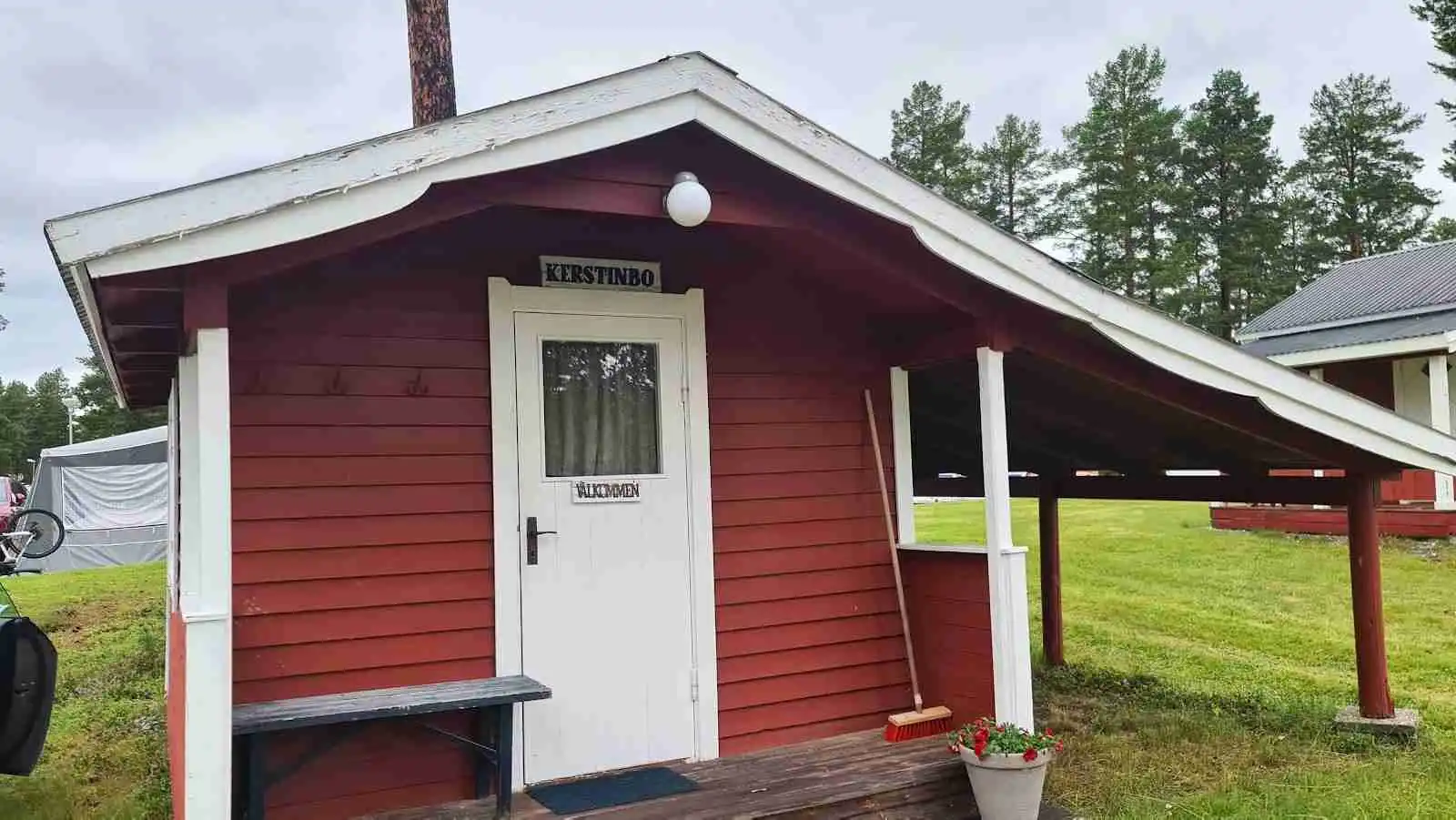 En trästuga med sidospår av trä, en dörr och ett skjul, omgiven av skuggade trädgårdsanläggningar och en trädgårdsyta på Vemdalens camping.