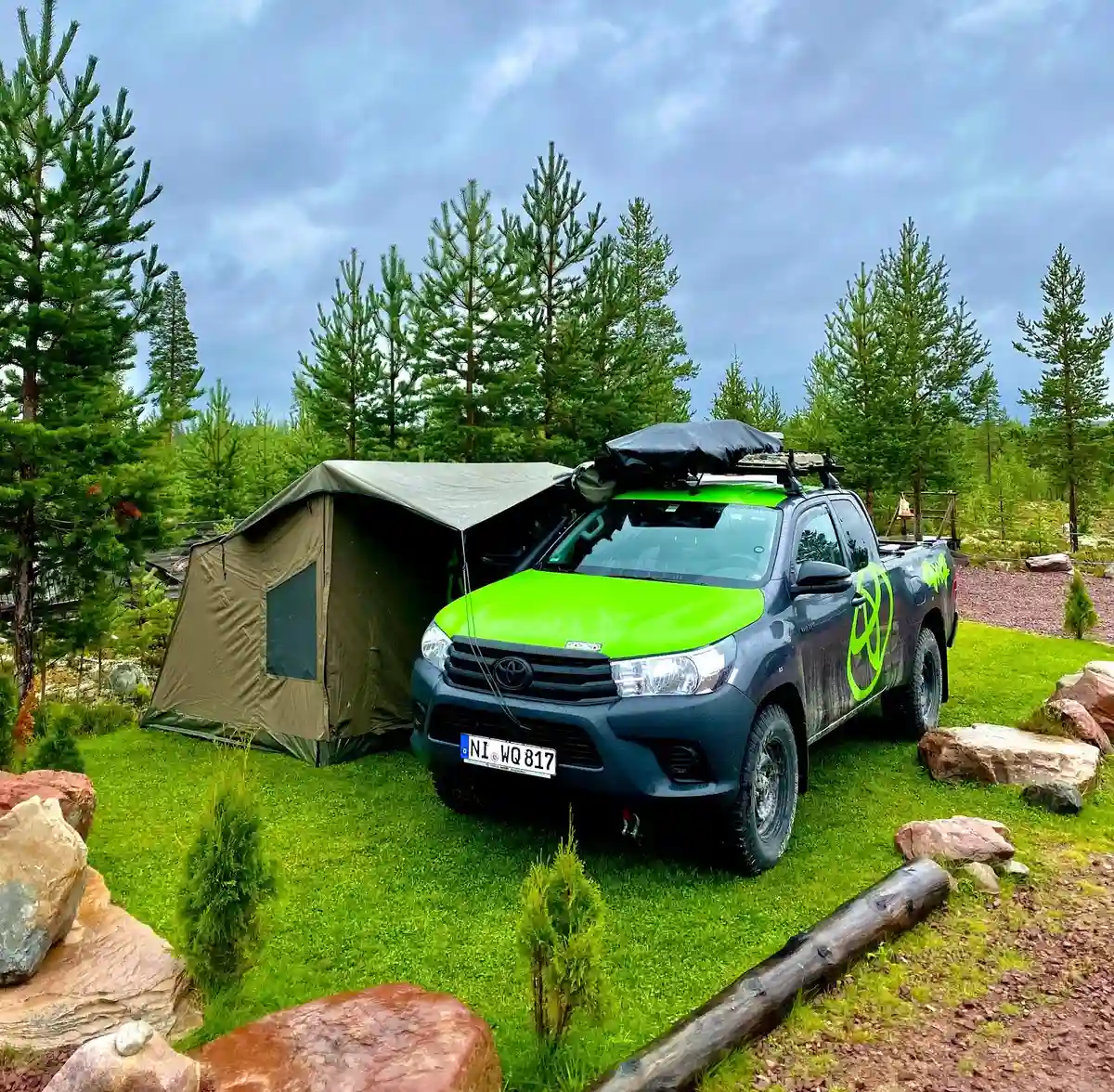 En pickuptruck utrustad med ett bagageräcke och terrängutrustning står parkerad i en naturskön alpbygd. I bakgrunden syns bergstoppar, vilket förstärker känslan av ett äventyrligt offroadcamp-besök.