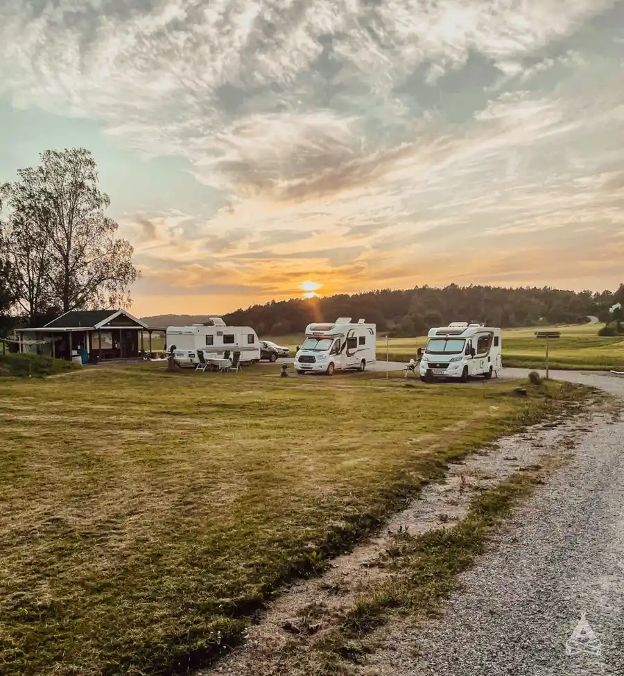 En campingplats vid Brålands Gård Nature Camp under skymningen, med en husvagn, en lastbil och ett tält uppställda på fältet i solnedgångens varma sken.