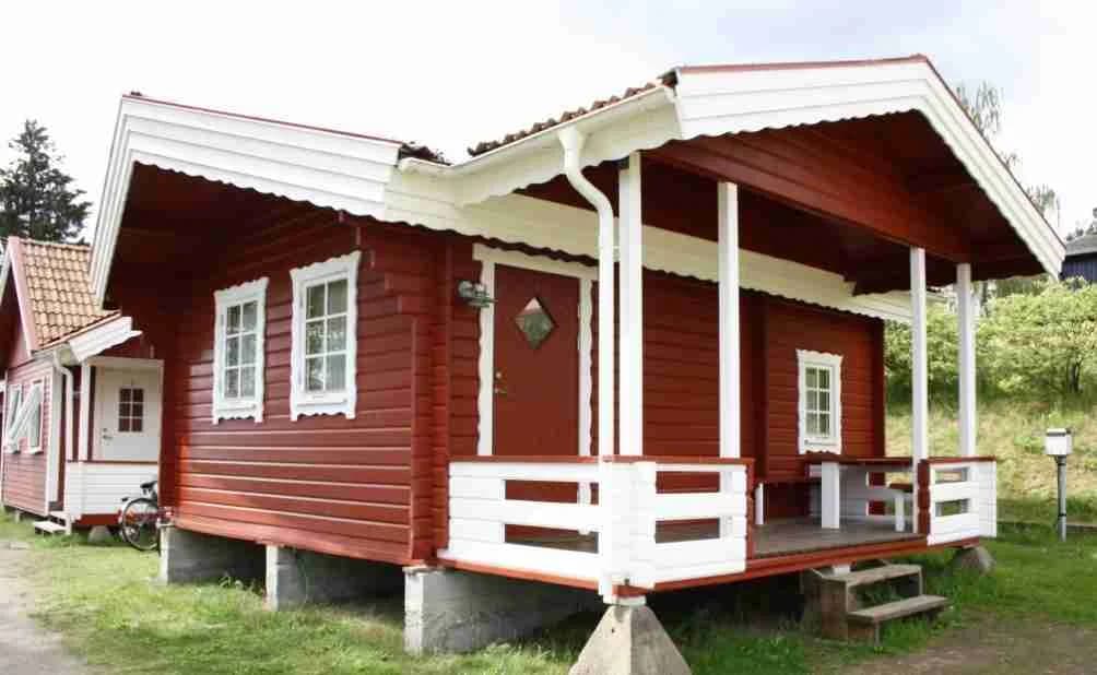 En liten trästuga med ett rött tak står på en grön gräsmatta vid Vegby Camping. Stugan har en veranda och omges av träd.