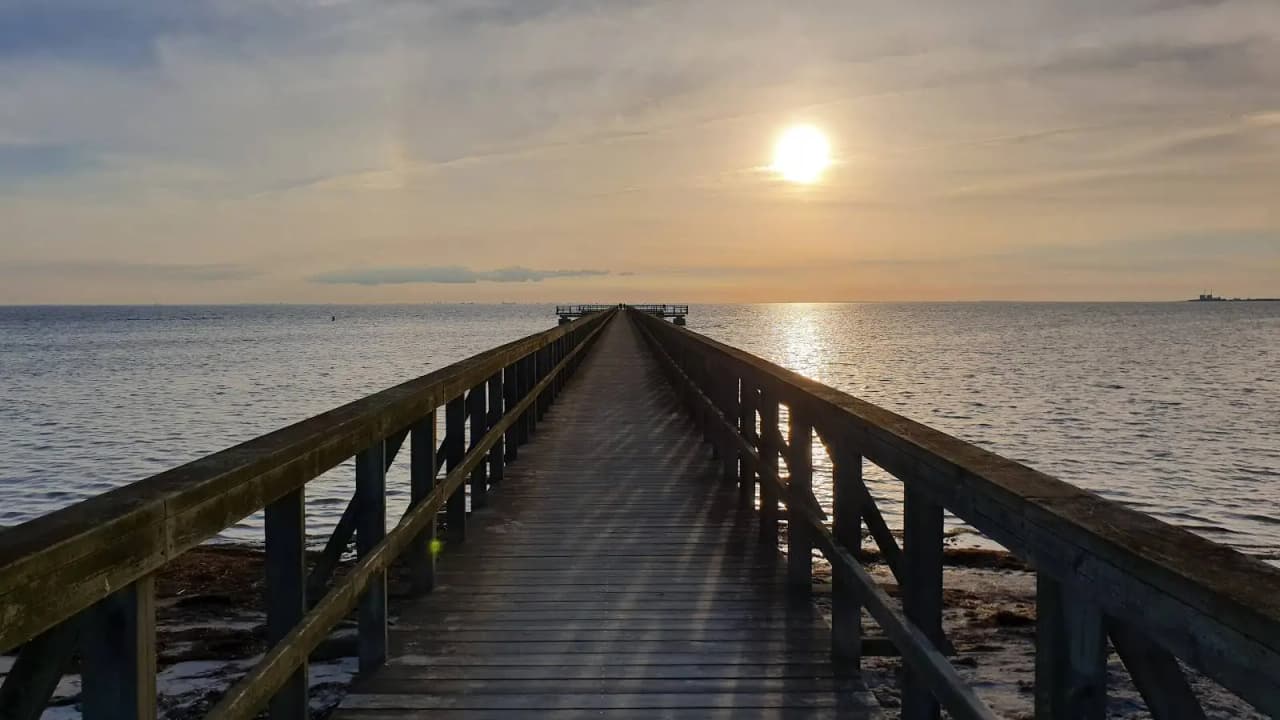 En solig dag vid Lomma Camping med blå himmel och en brygga som sträcker sig över det lugna vattnet.