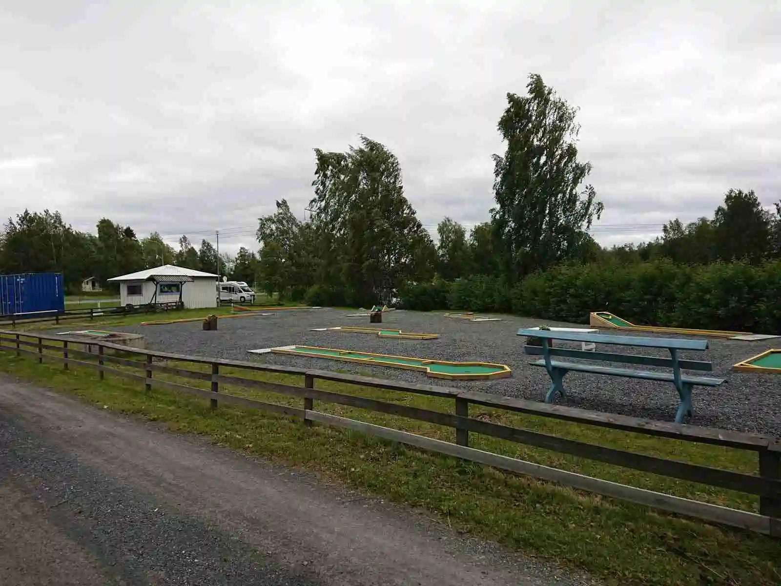 En öppen grön yta på Krokomvikens camping med en bänk i förgrunden, omgiven av gräsmatta och några trädstolpar, under en himmel med vita, fluffiga moln.