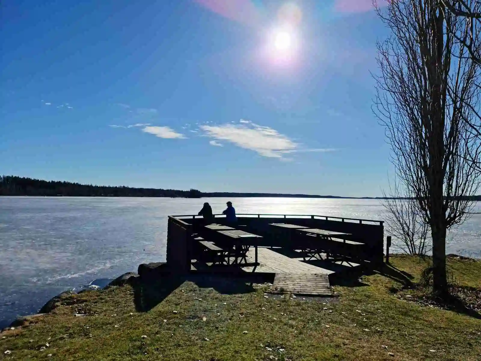 En bild av Vegby camping med en utomhusbänk vid en vattenkant. En klarblå himmel sträcker sig över horisonten, och vattnet glittrar i solljuset.