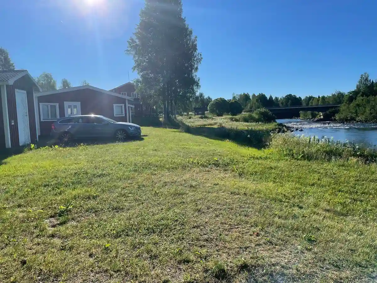 En grön och lummig campingplats i Fällfors med gräs och enstaka stugor, omgiven av ängsmark och landsbygdsmiljö.
