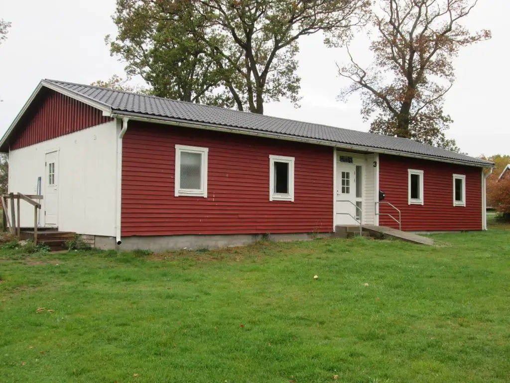 En röd trästuga med vita knutar omgiven av grönska på Kustgården Senoren, inkluderar gräsmatta och skog i bakgrunden.