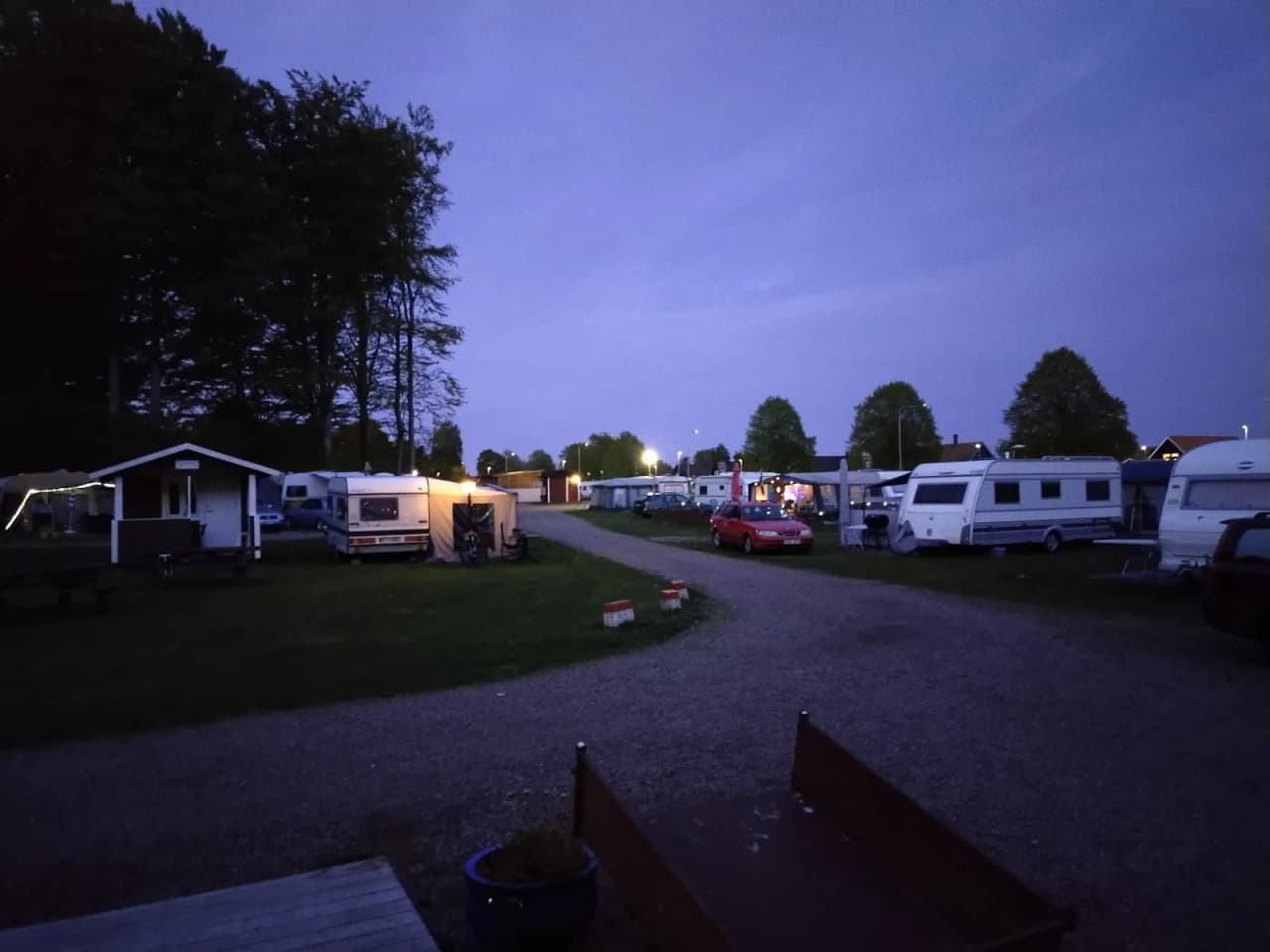 Bild på Elfdalens camping vid skymning. Flera husvagnar och husbilar står parkerade på rad på en grusplan, omgivna av träd. Ljuset från den nedgående solen skapar en stämningsfull atmosfär.
