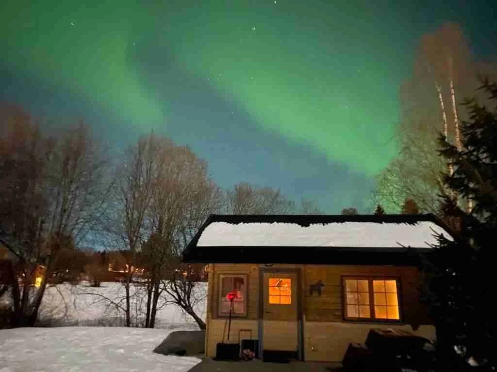 En stuga täckt av snö i ett vinterlandskap under natten, med norrsken synligt på himlen, vid Yttermalungs camping & stugby.