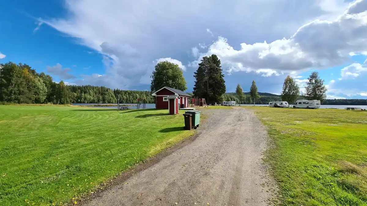 Bild av Vindelforsarnas camping: En grönskande gräsmatta sträcker sig mot ett litet rött stugområde, omgiven av öppen äng. Bakom stugorna syns en glittrande sjö eller reservoar, omgiven av pittoresk natur och parkmiljö.