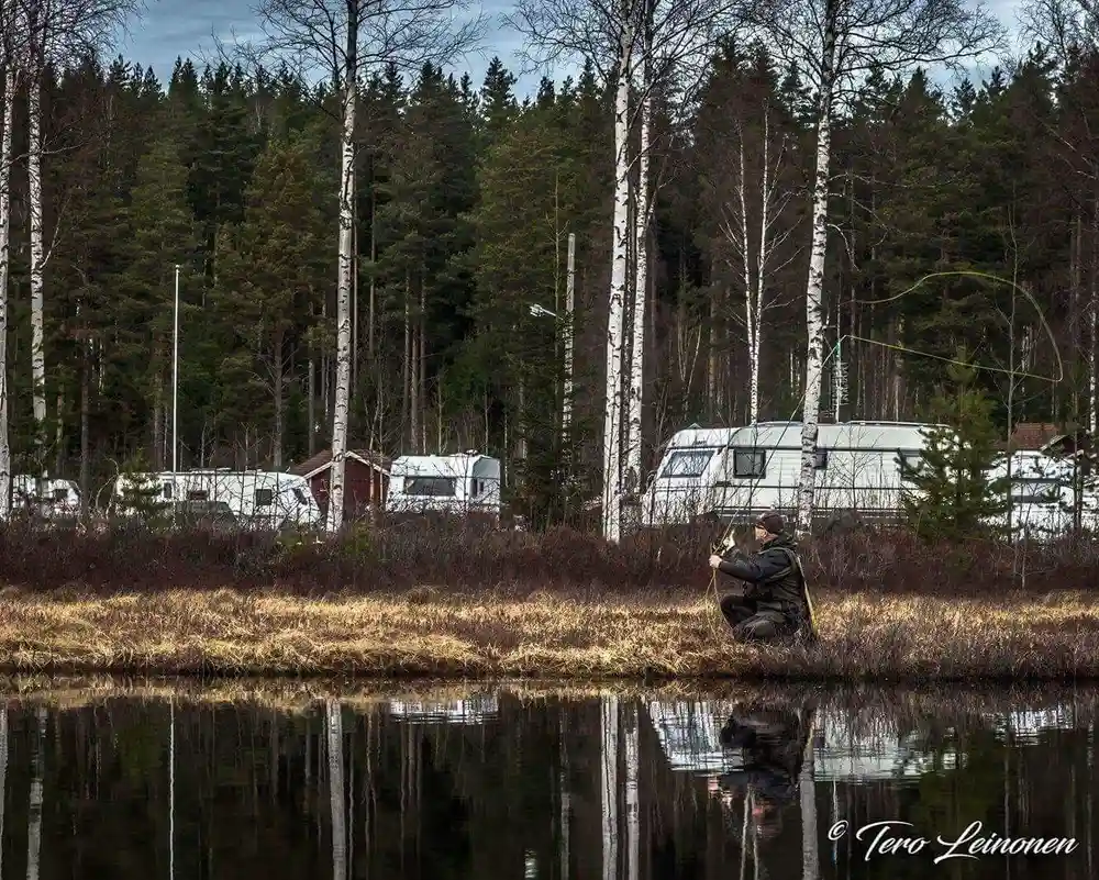 En husvagn står parkerad nära en stilla damm i ett våtmarksområde med skog i bakgrunden och reflekterande vattenytor i förgrunden.