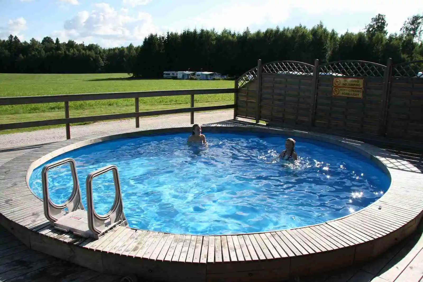 En camping med pool där människor njuter av fritidsaktiviteter och avslappning i solen. Utemöbler och parasoller finns runt poolen för skugga och komfort.