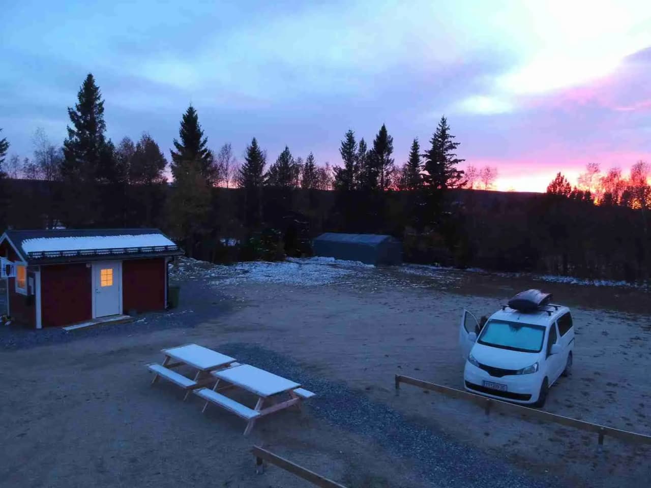 En vinterbild av Storsjö Camping och Ställplats, omgiven av snötäckta granar och med ett sken av skymning eller gryning i bakgrunden. Det finns små byggnader med frost på taken och is på marken.