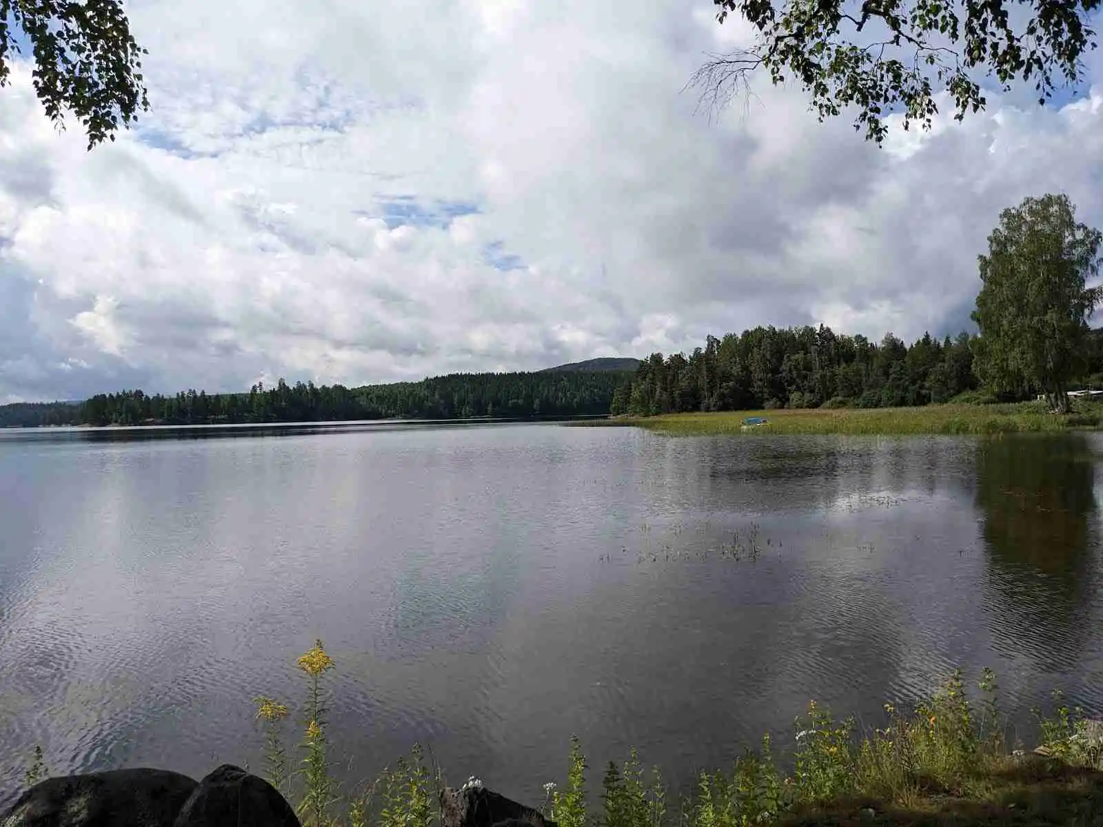 En naturskön campingplats vid en sjö med klarblått vatten och omgivande gröna träd under en himmel med lätta moln.