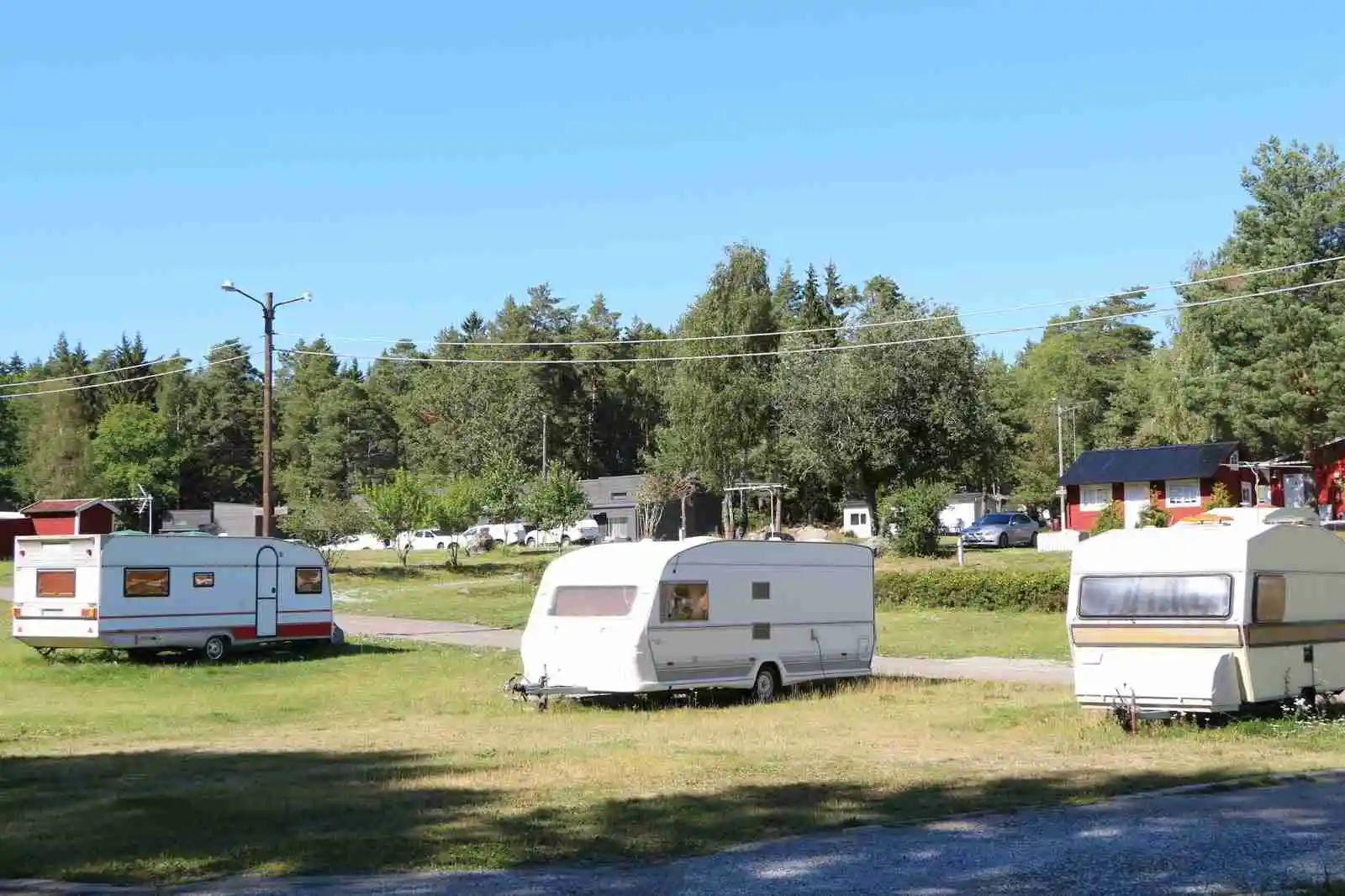 En campingplats med flera husvagnar och husbilar parkerade på en gräsyta. Flera träd i bakgrunden och en öppen parkmiljö syns tydligt.