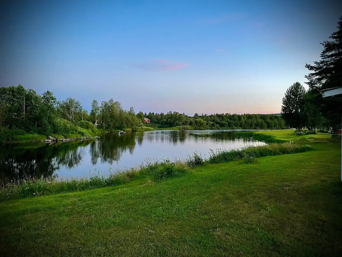 Bild av Fällfors Camping: En vidsträckt blå himmel speglas i det stilla vattnet, omringat av grönskande natur och träd som ramas in av den natursköna omgivningen.