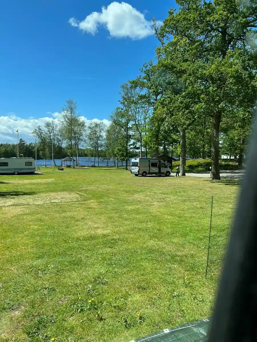 En grönskande campingplats i Alsterbro, med en välskött gräsmatta och skuggande träd. En husvagn är parkerad vid sidan, och cumulusmoln syns på himlen.