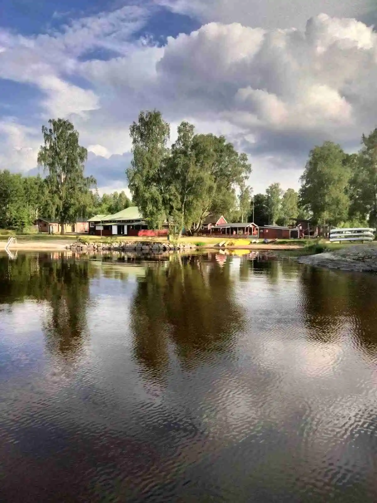 En naturskön sommarbild av Hätteboda vildmarkscamping med lugnt vatten som reflekterar omgivande träd och växtlighet.