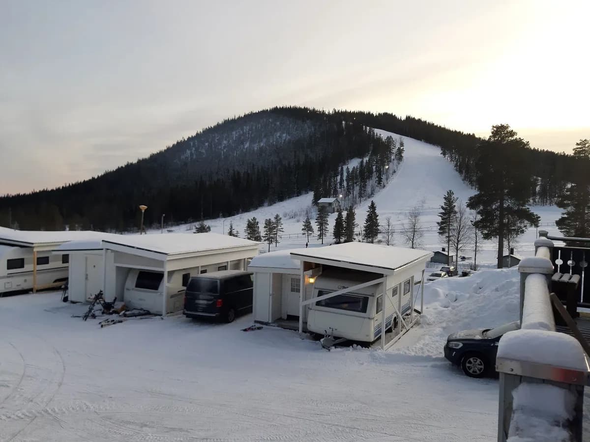 En vinterbild av Storklinta Skidcenter med snöklädda tallar och lärkträd. En stuga står i bakgrunden, och en husbil är parkerad framför, täckt av snö.