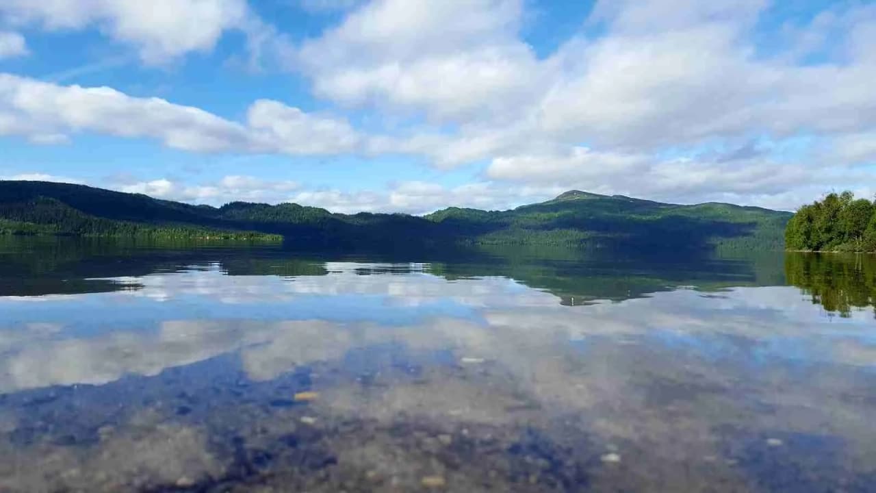En vidsträckt vy över Lia Ranch vildmarkscamping med en klarblå himmel fylld med moln, en spegelblank vattenyta i förgrunden och en naturlig horisontlinje i fjärran.